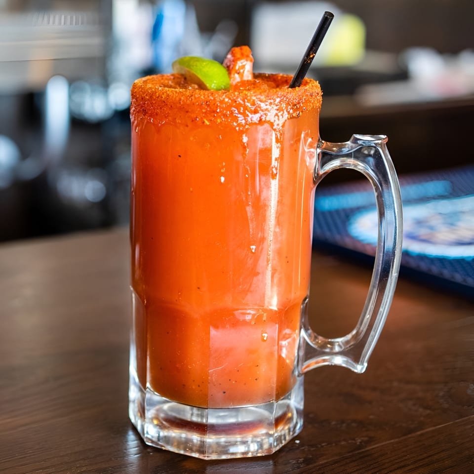 A large glass mug of Michelada, garnished with a lime wedge, shrimp, and a black straw.