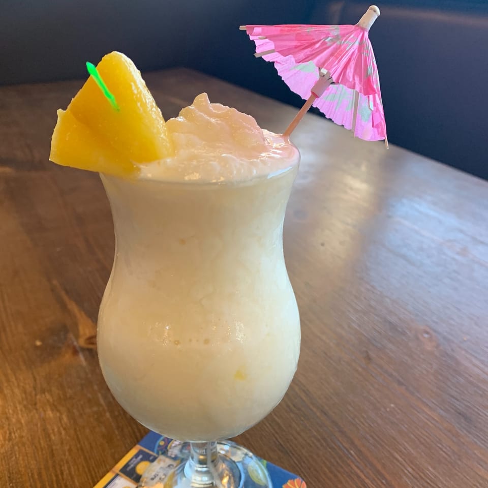 Creamy frozen drink with a pineapple wedge and pink umbrella on a wooden table.