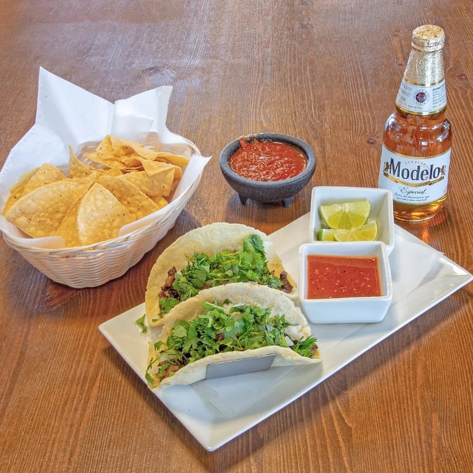 Mexican tacos with cilantro, chips, salsa, lime wedges, and a Modelo Especial beer on a wooden table.