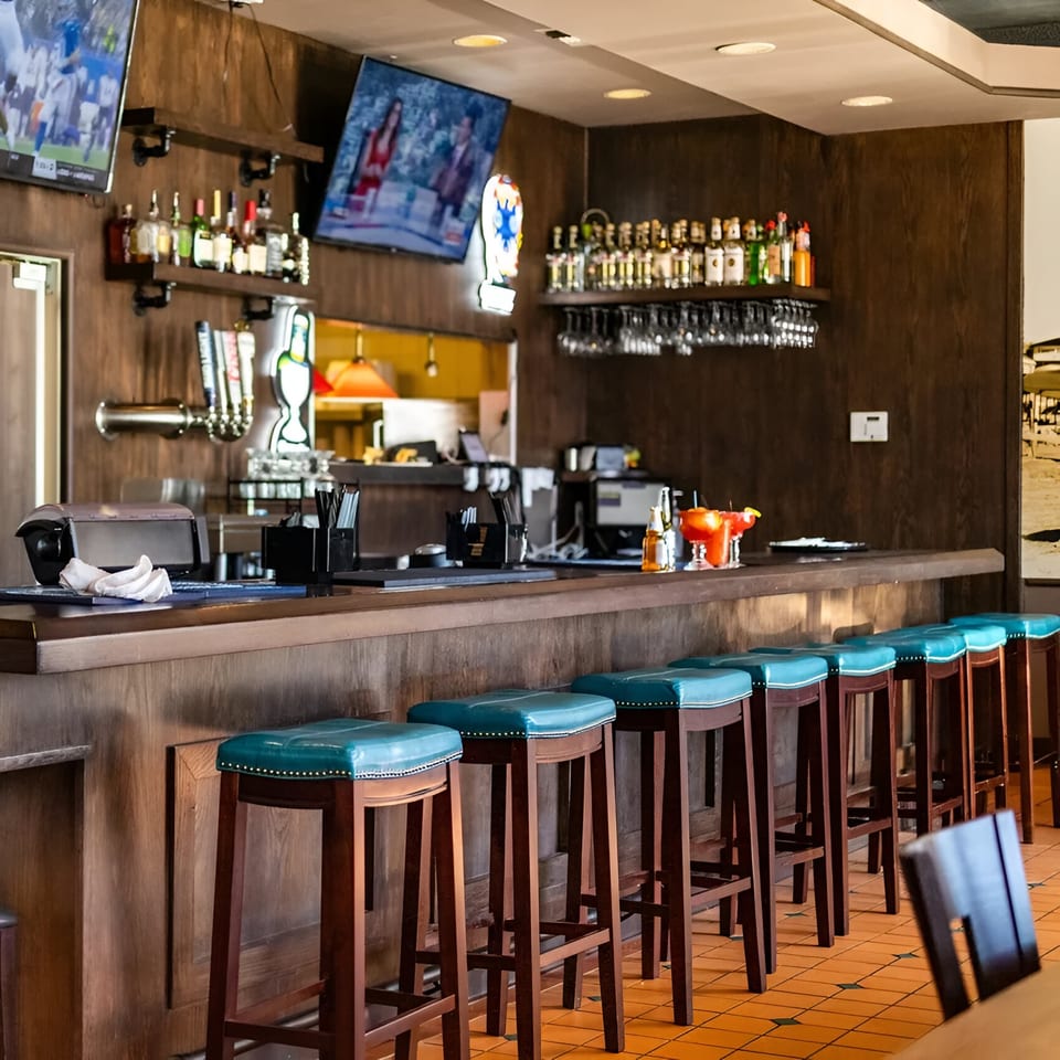 A long wooden bar with teal stools, liquor bottles, and two TVs showing sports.