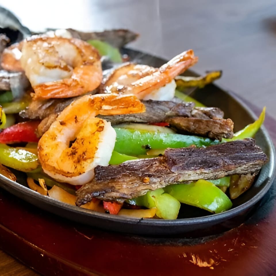 Sizzling fajitas with shrimp, steak, green and red bell peppers, and onions on a dark platter.