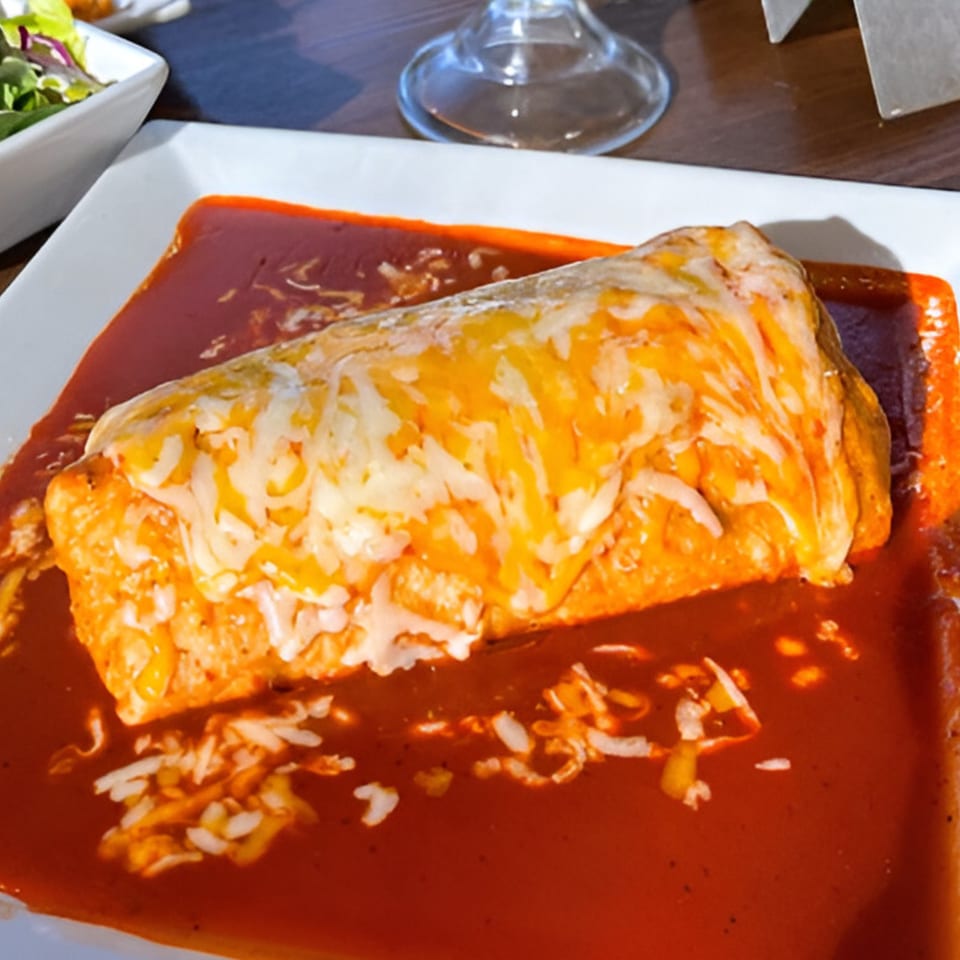 A large, cheese-covered burrito smothered in red sauce on a white plate at Ruben's Mariscos.