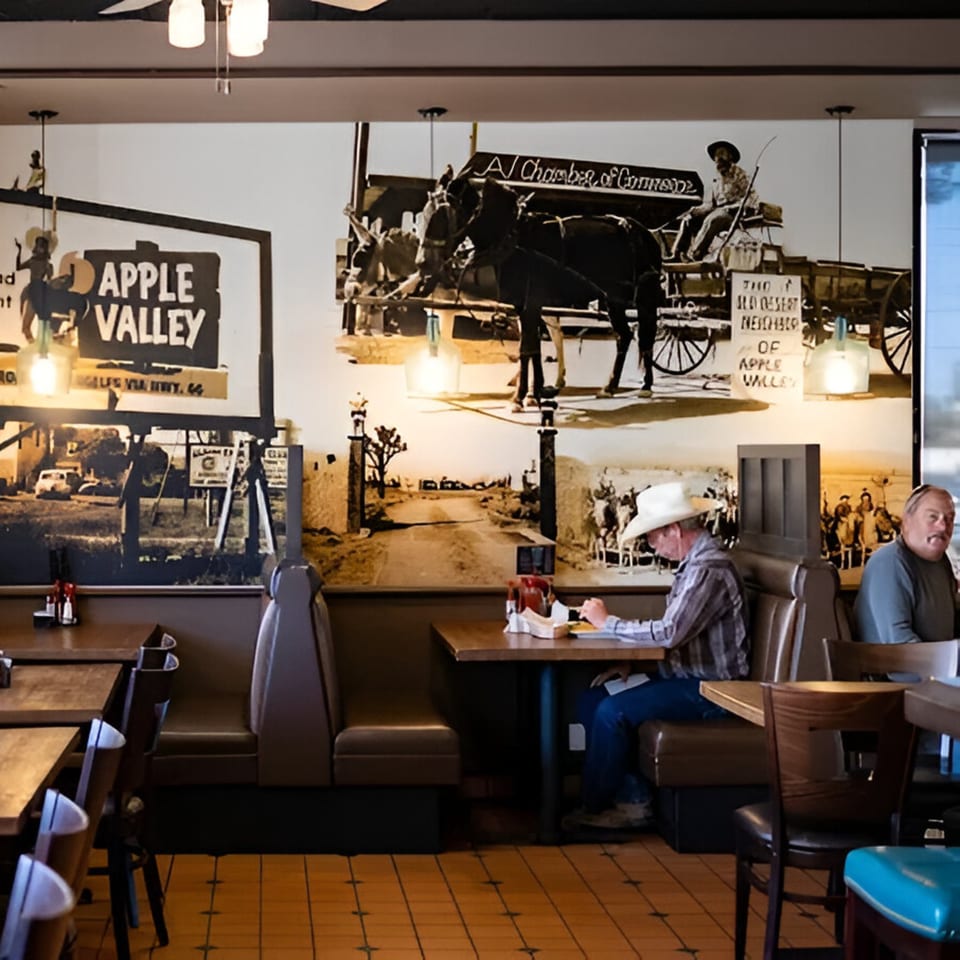 Restaurant interior with patrons dining at tables, featuring a large mural of vintage Apple Valley scenes.