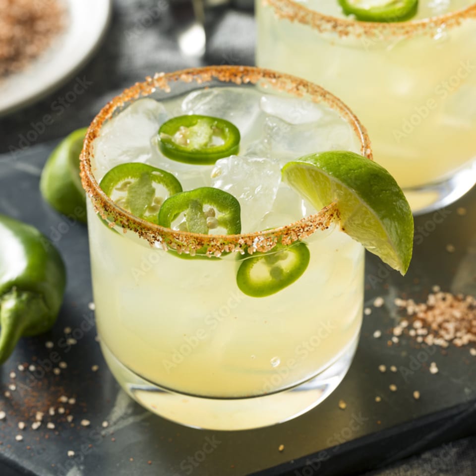 Spicy margarita with jalapeño slices, lime wedge, and a chili-salt rim on a dark surface.