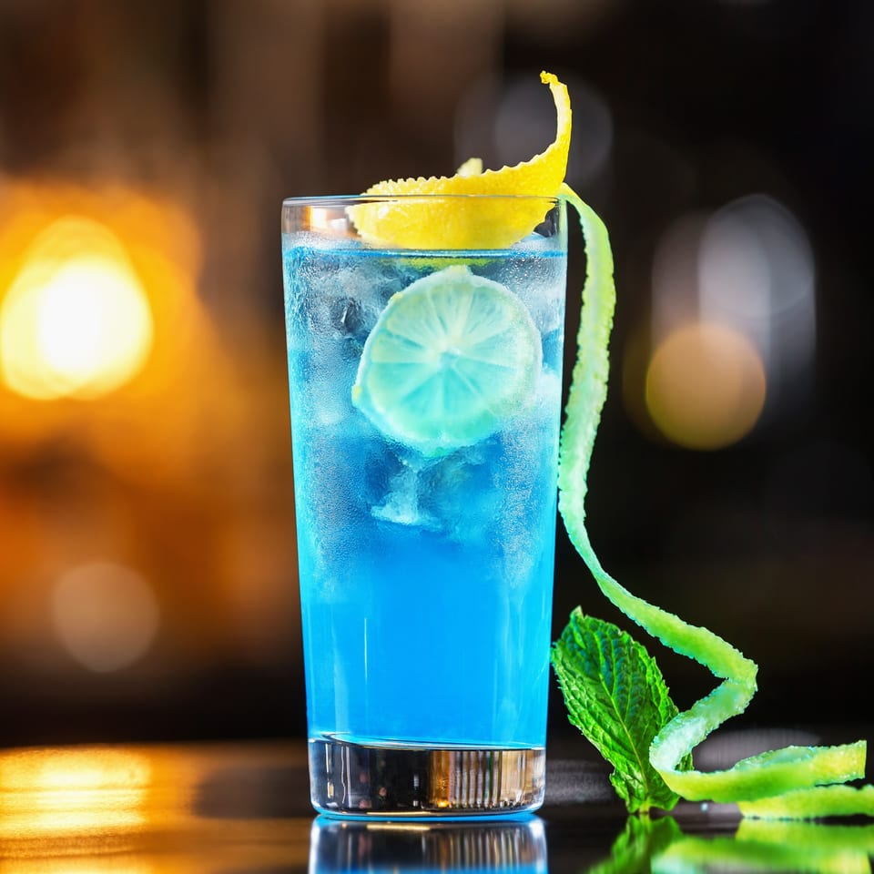 Vibrant blue cocktail with a lime slice, lemon peel, and mint leaf on a dark, blurred background.