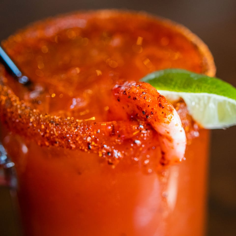 A close-up of a Michelada cocktail with a chili-lime rim, shrimp, and a lime wedge.