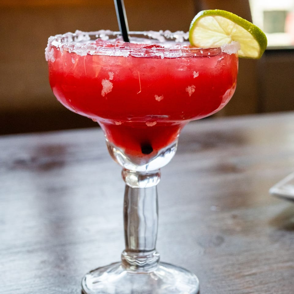 A vibrant red margarita with a salted rim, lime wedge, and straw on a wooden table.