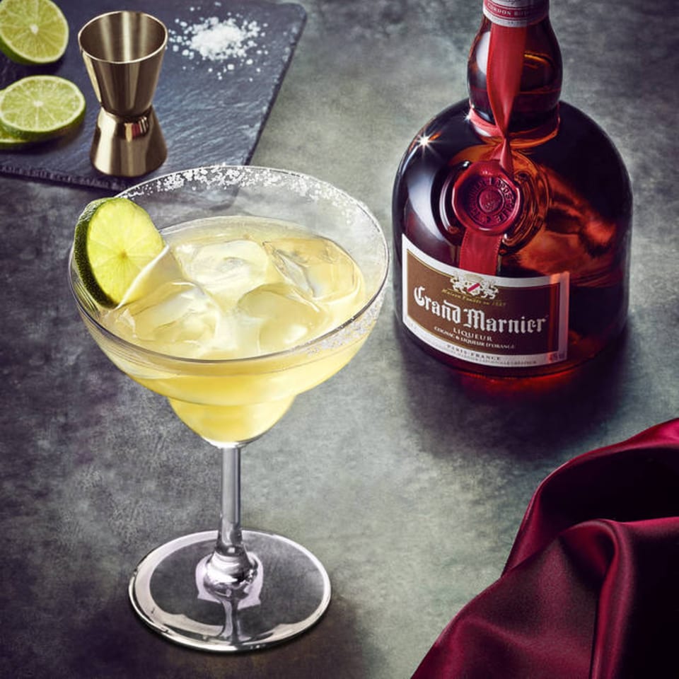 Margarita with lime and salt, Grand Marnier bottle, jigger, and lime slices on a dark surface.